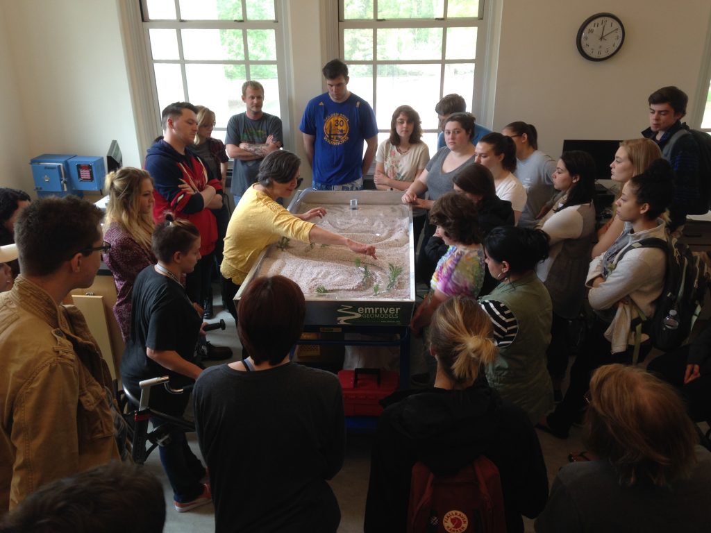 Dr. Jackie Gallagher demonstrates lateral erosion and fluvial hazards using the stream table while students look on.