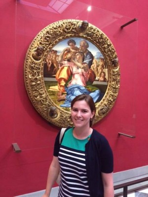 Photo shows woman standing in front of red wall with circular, gold-framed piece of art hanging behind her