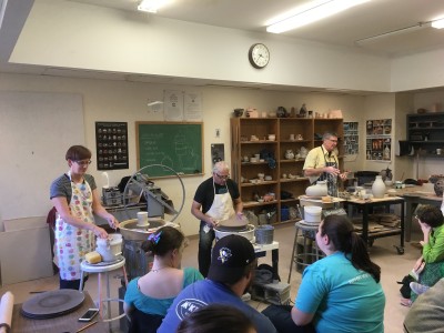 image shows three artists throwing pottery on pottery wheels while students sit and observe