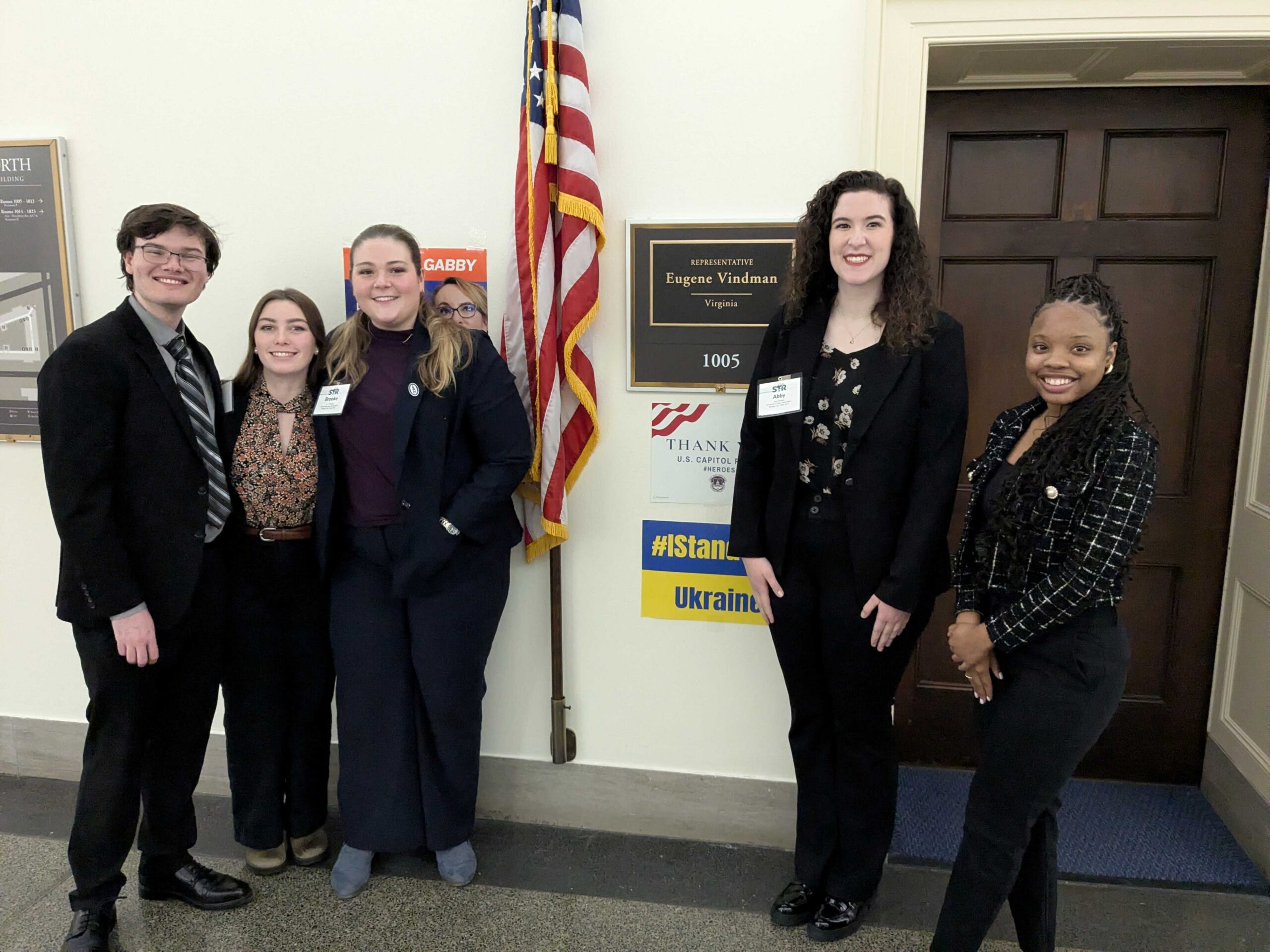 Students and congressional aide pose in front of Rep. Eugene Vindman's capitol hill office door with a US flag in the background,