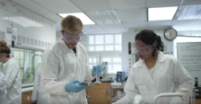 Two students in goggles and laboratory coats working on an experiment in a laboratory.