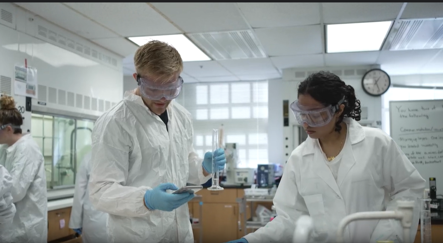 Two students in goggles and laboratory coats working on an experiment in a laboratory.