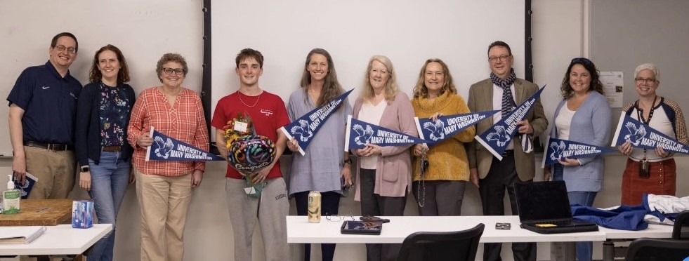 A group of faculty with a student standing in front of a classroom. The student is being congratulated for winning the Goldwater scholarship. The faculty are holding UMW pennants.