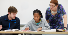 Two students looking at textbook and working on solving problems with a faculty member observing. 