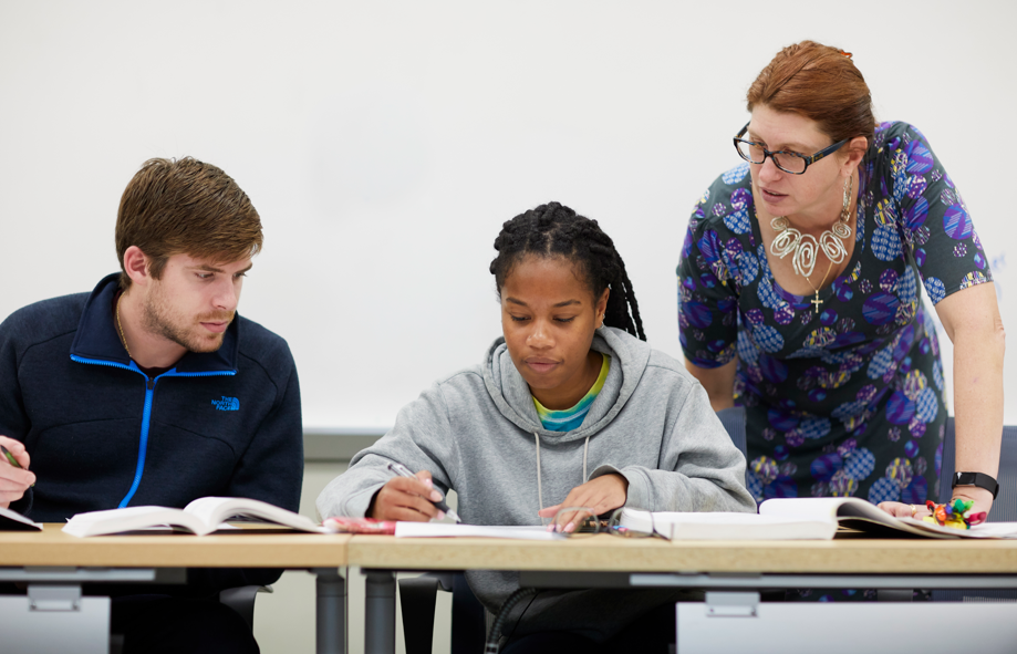 Two students looking at textbook and working on solving problems with a faculty member observing. 