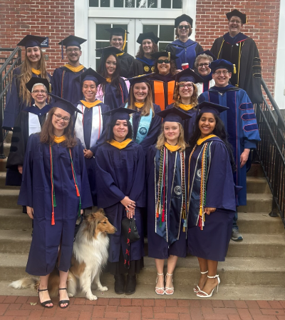 Graduates and faculty dressed in caps and gowns before commencement. Individuals are posed on steps in front of a building.