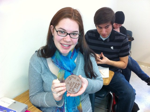 A young woman seated at a desk holds a hand-sized clay tablet with inscriptions on it. 
