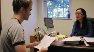 Professor Lee sits at a desk and talks with a student across from the desk and who holds a printout. 