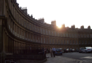 An image of a curving building row with cars parked along the street, people talking, and sun rays peeking over the top of the building.