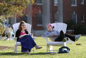 Two students sit in Adirondack chairs on campus, reading and talking.