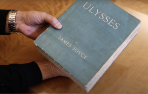 Barrenechea's hands hold a copy of a blue hardcover book. The top of the cover has capitalized gold letters identifying the title Ulysses, while the bottom of the cover has James Joyce. No other images appear.  
