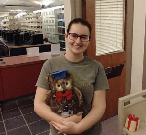 A smiling Madison Roberts poses with the Simpson OWL with library book shelves in the background.