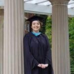 Kaitlie Goodwin stands in graduation regalia between two architectural columns.