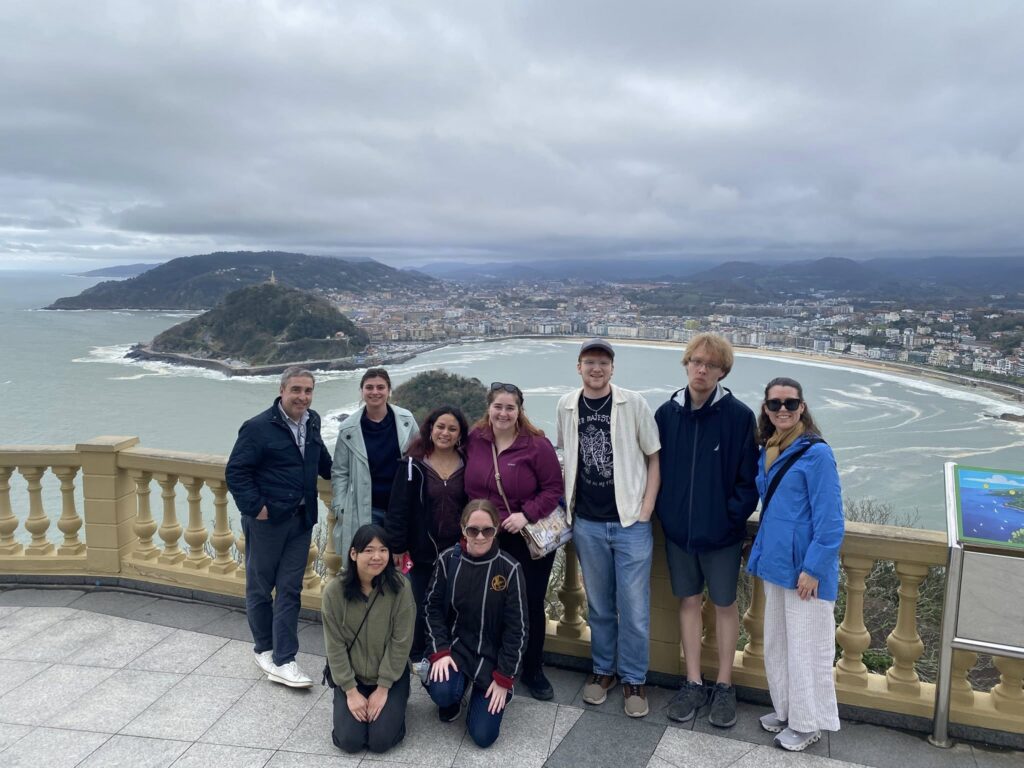 A group of students and faculty standing in front of a railing outside. Behind them is a cityscape with a bay and mountains surrounding it.
