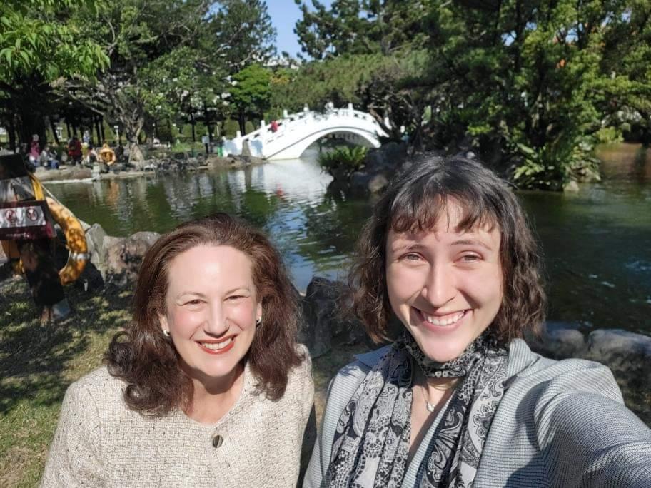 Tess Darroch with Professor Elizabeth Larus in Taiwan. In the background is pictured lots of nature including trees, a small body of water, and a bridge across the water. Some other people are in the far background as well.