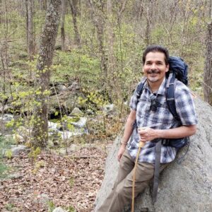 Professor Ranjit Singh on a hiking trip seated on a large rock in a forest.