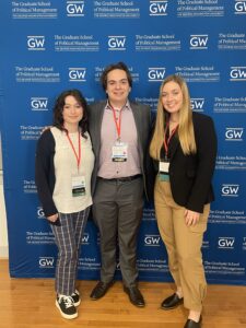 Sasha, Sam, and Abby posing for a picture with a photo backdrop that says The George Washington University: The Graduate School of Political Management.