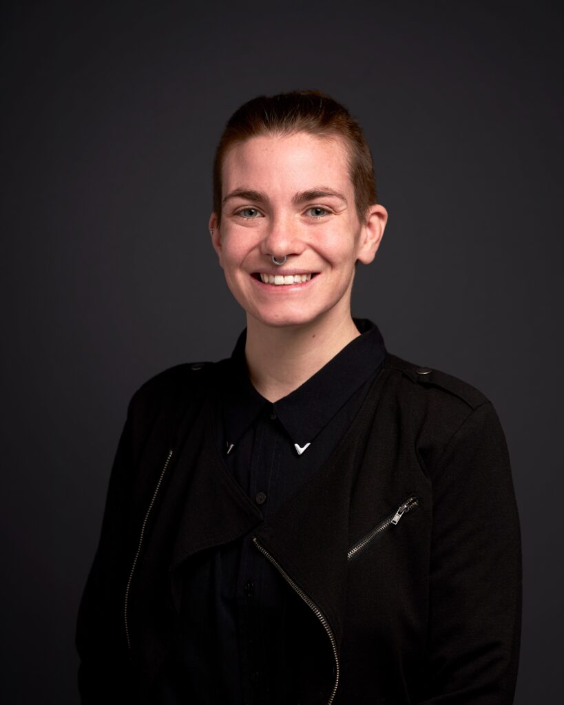 A person with short hair and a septum piercing smiling, wearing dark clothes and photographed against a black background.