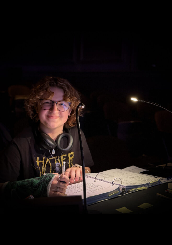 A theater student sits at a stage management console, smiling warmly while working on a production.
The student wears glasses, a black t-shirt with yellow graphic text, and a pair of large black headphones around their neck. They are holding a pencil, poised to make notes in a large, open white three-ring binder that appears to be a prompt book or script. The workspace is illuminated by two small, flexible gooseneck task lights in an otherwise dark theater setting.