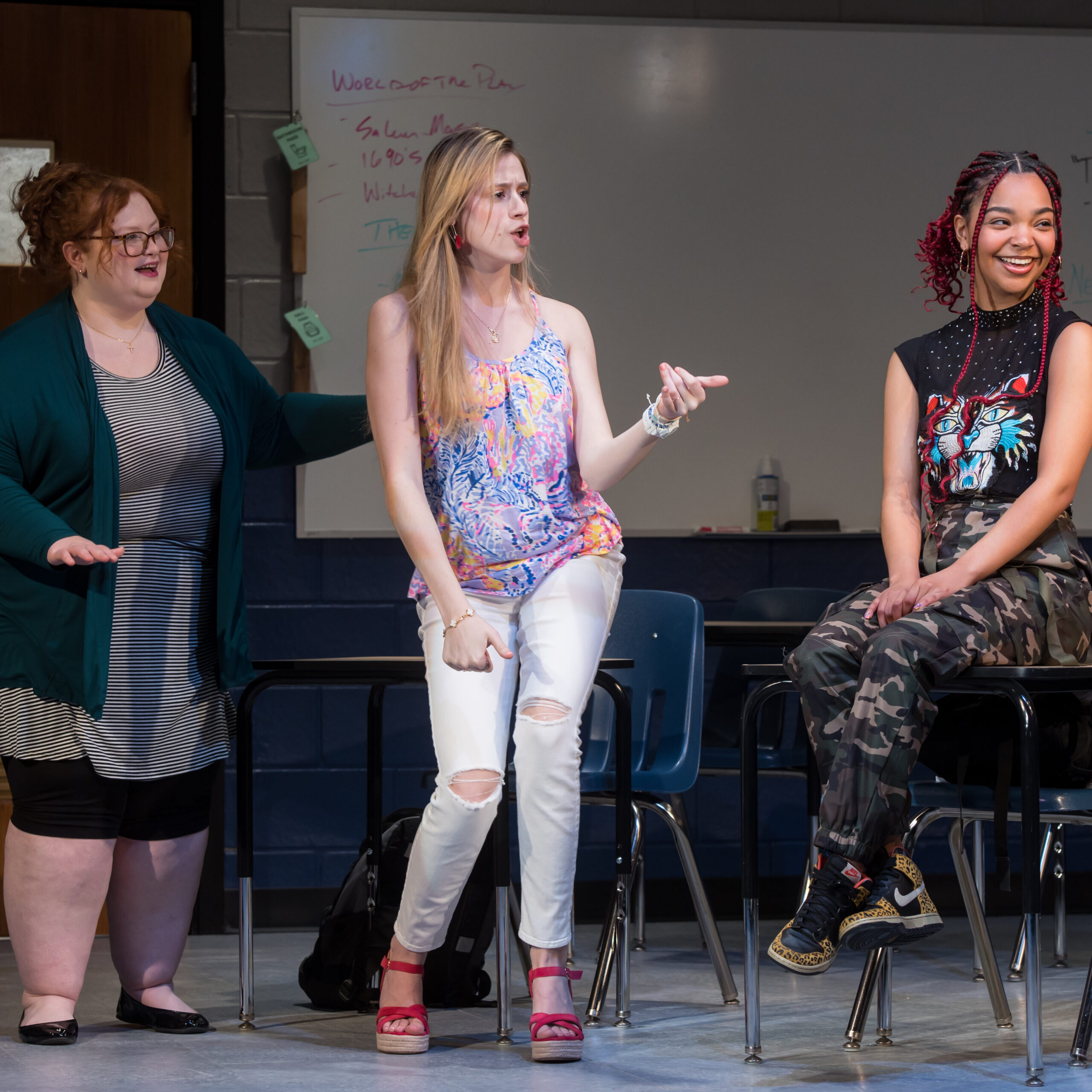 The central figure is standing with a surprised expression, mouth open, wearing a purple and orange patterned sleeveless top, white ripped jeans, and red platform sandals. To her right, another woman sits on a high stool, smiling; she has bright red braided hair and is wearing a black graphic t-shirt and camouflage pants. On the left, a third woman stands wearing glasses, a striped top under a deep green cardigan, and black leggings.