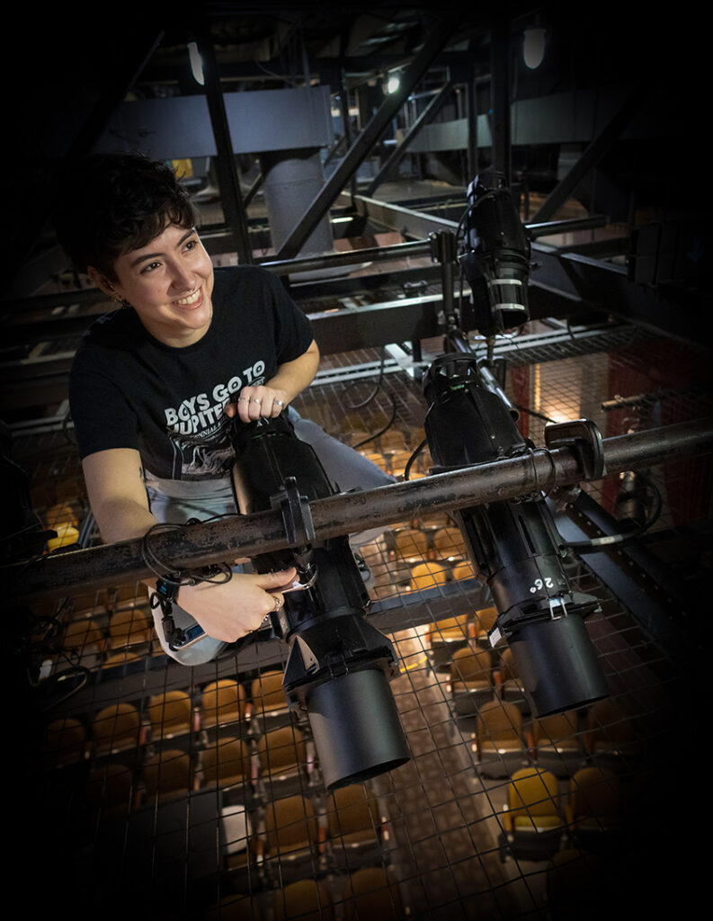 A technical theatre student at the University of Mary Washington's Klein Theatre smiles while adjusting large black spotlights from a high lighting rig. The student wears a black t-shirt featuring the phrase