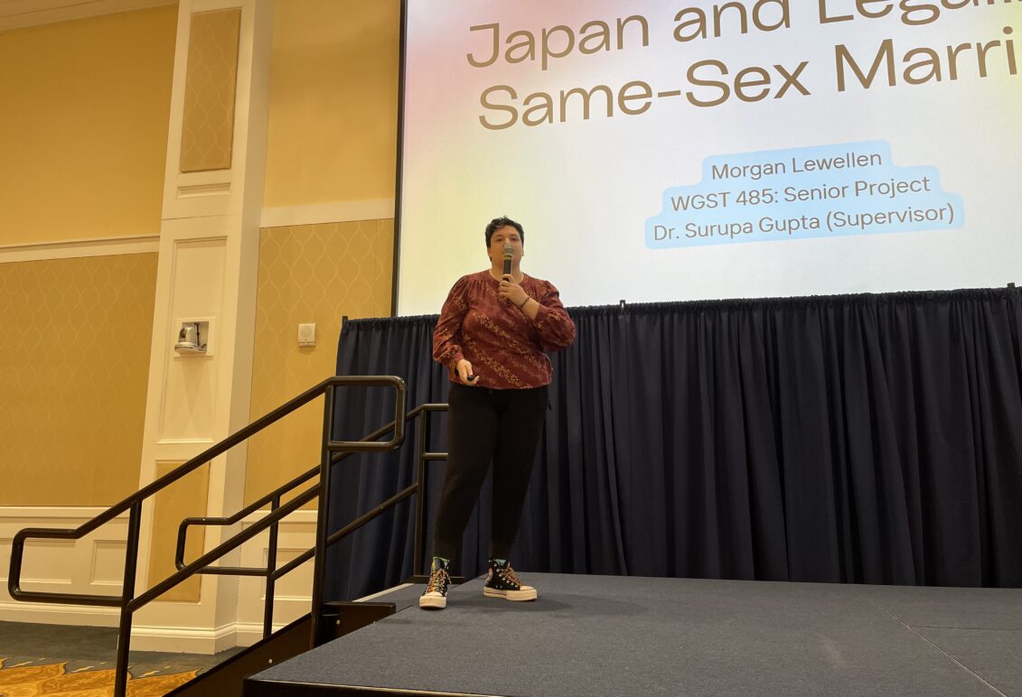 A person holding a mic presenting in front of a projected slideshow presentation which states "Japan and legalizing same-sex marriage." 